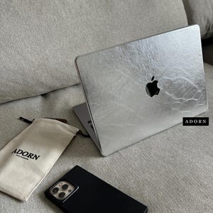 Puede incluir: Una pegatina metálica plateada con acabado texturizado aplicada a un portátil, con el logotipo de Apple recortado. Junto a él hay una funda de smartphone negra y una bolsa de tela beige con el texto ADORN impreso en negro.