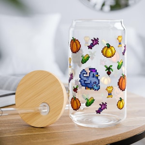 Może przedstawiać: Przezroczysta szklanka w kształcie puszki z wzorem pixel art przedstawiającym dynie, kukurydzę, buraki, pszenicę i niebieskiego kurczaka. Zestaw zawiera okrągłą bambusową pokrywkę z otworem na słomkę oraz szklaną słomkę do napojów.