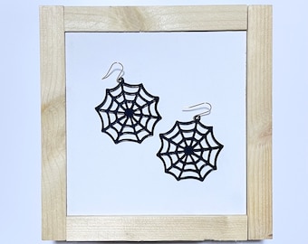 May include: A pair of black spiderweb earrings with silver-colored hooks. The earrings are a Halloween-themed accessory, featuring a detailed web design. The earrings are displayed on a white background within a wooden frame.