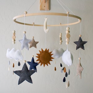 May include: A wooden baby mobile with a white, gray, and brown color scheme. The mobile features a sun, moon, clouds, stars, and wooden beads.