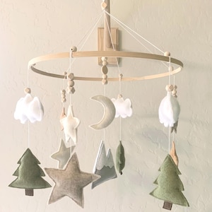 May include: A wooden baby mobile with felt woodland animal and nature-themed decorations, including clouds, stars, a crescent moon, mountains, and pine trees. The mobile is suspended from a wooden ring and hangs from a wooden arm.