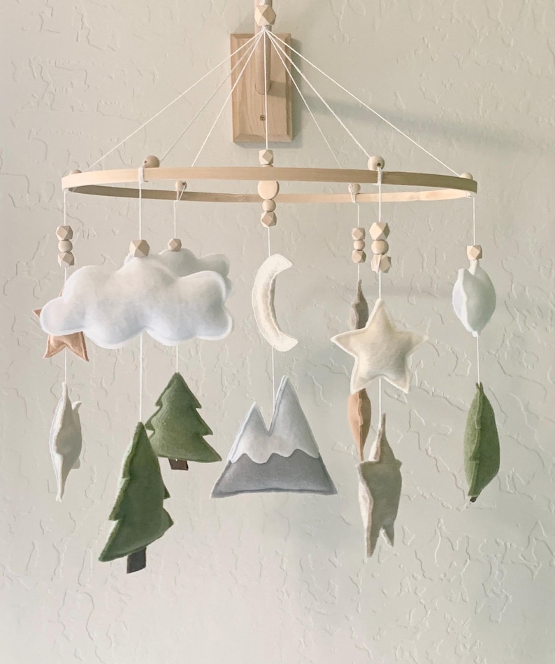 May include: A wooden baby mobile with felt woodland animal and nature-themed decorations. The mobile features a cloud, moon, star, mountains, and trees.