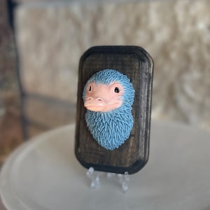 Op de afbeelding: Een houten plaquette met een blauw en roze wezen met een getextureerd, veervormig uiterlijk. De kop van het wezen is gecentreerd op een donkerbruine, rechthoekige plaquette. De plaquette wordt gepresenteerd op een wit bord.