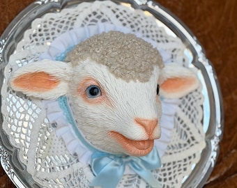May include: A sculpted lamb head wall plaque mounted on a round silver-toned tray. The lamb has textured beige wool, white fur, and blue eyes. It is framed by a white lace doily and a white ruffled collar, accented with a light blue satin bow.