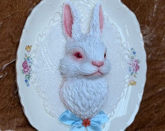 Op de afbeelding: Een witte konijnenkopsculptuur met rode ogen en roze binnenoren, gemonteerd op een ovaal keramisch bord. Het bord heeft een bloemenrand, een witte kanten achtergrond en een lichtblauwe satijnen strik aan de onderkant van het konijn.