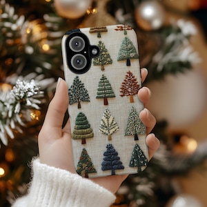 May include: A phone case featuring a repeating pattern of embroidered Christmas trees in various colors, including green, brown, and blue, on a light beige background. The case has a black border and a camera cutout. The case is held in a person's hand.