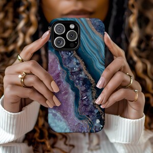 May include: A smartphone case with a marbled design in shades of blue, purple, and gold. The case has a glossy finish and a black border. The phone case is held in front of a person wearing a white sweater.