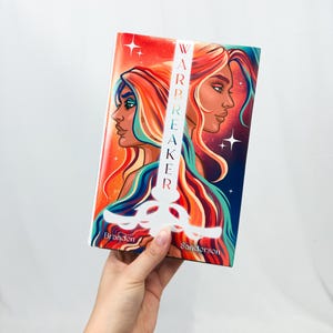 Puede incluir: Un libro de bolsillo titulado "Warbreaker" de Brandon Sanderson. La portada presenta dos figuras femeninas estilizadas con cabello ondeante en tonos de rojo, naranja y azul. El título está alineado verticalmente en blanco, con un diseño de espada.