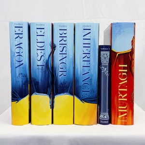 Puede incluir: Una colección de novelas de fantasía de Paolini. Las lomos muestran títulos como "Eragon", "Eldest", "Brisingr", "Inheritance" y "Murtagh". Los libros tienen cubiertas azules y amarillas con ilustraciones y texto estilizados. También hay un libro más pequeño con el lomo azul oscuro.