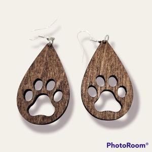 May include: A pair of brown wooden teardrop-shaped earrings with a paw print cutout. The earrings have silver hooks.