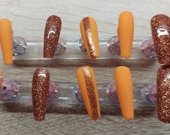 Peut inclure: Ensemble de dix faux ongles longs, orange et pailletés cuivre. Les ongles sont disposés en deux rangées sur un présentoir transparent.