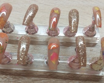Peut inclure: Dix faux ongles présentés sur un support en plastique transparent. Les ongles sont disposés en deux rangées de cinq. Les ongles sont nus avec des motifs de feuilles d'automne et des paillettes dorées.