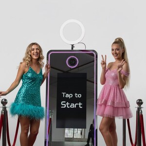 May include: A photo booth with a black mirrored surface and a pink frame. The screen displays "Tap to Start". Two people stand on either side of the booth, one in a teal sequined dress with feather trim, the other in a pink sequined dress.