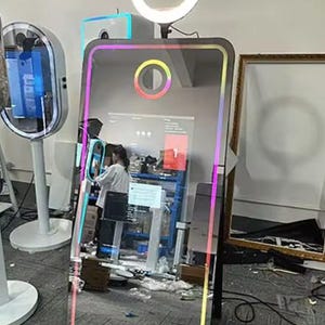 May include: A large, rectangular photo booth mirror with a colourful LED border. The mirror has a circular light and a smaller circular camera. A person is visible in the reflection, operating the photo booth. Other photo booths are in the background.