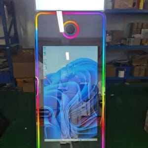 May include: A tall, rectangular photo booth with a vibrant, multi-coloured LED border. The booth has a large screen displaying a blue and white abstract image. A bright white light panel is at the top.