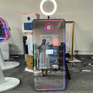 May include: A large, rectangular photo booth with a reflective surface and a colourful LED border. A circular light is mounted above the booth. The booth is in a room with other equipment and a person is reflected in the mirror.