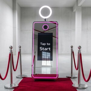May include: A contemporary photo booth with a large mirror, a ring light, and the text "Tap to Start" on the screen. The booth is framed with a pink neon light and sits on a red carpet. Silver stanchions with red velvet ropes are on either side.