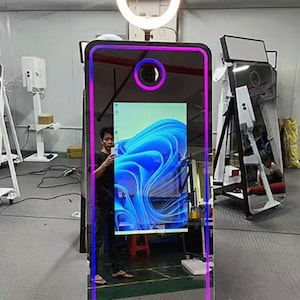 May include: A large, rectangular photo booth with a mirrored surface and a neon pink and blue border. The booth has a circular light at the top and a camera lens. The screen displays a blue and white abstract design.