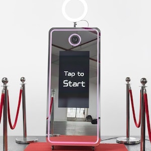 May include: A modern photo booth with a large mirror and a ring light. The screen displays "Tap to Start". It's set up on a red carpet, with velvet ropes and stanchions creating a pathway.