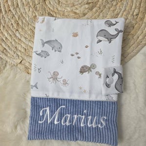 Könnte beinhalten: Eine Babydecke mit maritimem Thema, die Wale, Delfine, Schildkröten und Oktopusse zeigt. Der untere Teil ist aus blauem Cord, bestickt mit dem Namen "Marius" in Weiß.