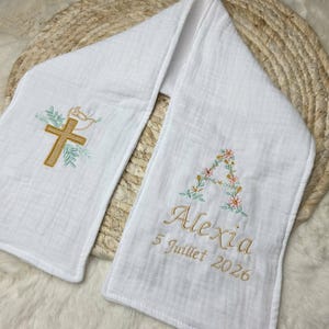 May include: White baptismal scarf with embroidered gold cross, dove, and floral accents. The scarf is personalized with the name "Alexia" and the date "5 Juillet 2026" in gold thread. The scarf is made of a soft, textured fabric.
