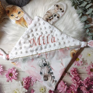 May include: A white baby security blanket with a floral print and the name "Mila" embroidered in pink. The blanket features animal-themed ears, including a lion and meerkat. The blanket is surrounded by pink flowers.
