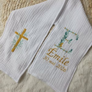 May include: White, textured scarf with gold embroidered cross and floral accents. The scarf features the name "Emile" and the date "30 mai 2026" in gold embroidery, along with a floral initial "E".