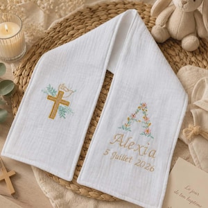 Peut inclure: Un tissu en mousseline blanc avec des broderies. Un côté présente une croix dorée avec des feuilles vertes et une colombe. L'autre côté affiche une lettre A florale, le prénom Alexia et la date 5 Juillet 2026 brodés en fil doré.