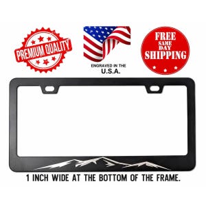 May include: A black rectangular license plate frame featuring a silver mountain range graphic along the bottom edge. The frame has two mounting holes at the top and is 1 inch wide at the bottom. The design is engraved in the U.S.A.