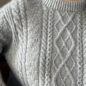 Może przedstawiać: Jasnoszary sweter z długim rękawem o teksturowanym splocie. Wzór obejmuje pionowe kolumny warkoczowe przeplatane sekcjami o splocie koszykowym oraz prążkowany dekolt wykończony ściągaczem.