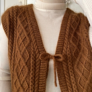 May include: A sleeveless, V-neck knit vest in a rich tobacco brown color, featuring a textured diamond cable knit pattern throughout. The vest is secured at the center front with a matching brown yarn tie fashioned into a small bow.