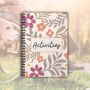 以下が含まれることがあります： 白い螺旋綴じのノート。花柄で、黒い枠の中に「アクティビティ」の文字が書かれている。花はピンク、オレンジ、黄色です。