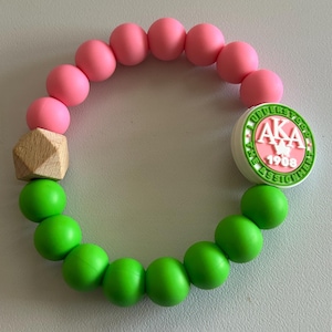 May include: A beaded bracelet featuring pink and green round beads, a wooden geometric bead, and a circular charm with the text "AKA 1908 UNDERSTOOD THE ASSIGNMENT" in pink and green. The bracelet is on a light gray surface.