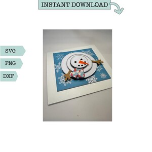 May include: A white snowman with a red nose and a blue and white plaid scarf is layered on top of a blue background with white snowflakes. The snowman is on a white card.