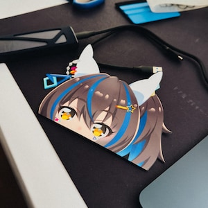 May include: A cut-out sticker featuring an anime-style character with brown hair, blue highlights, and large yellow eyes. The sticker has white cat ears and a star-shaped hair accessory. The background includes a black surface, scissors, and a blue tool.