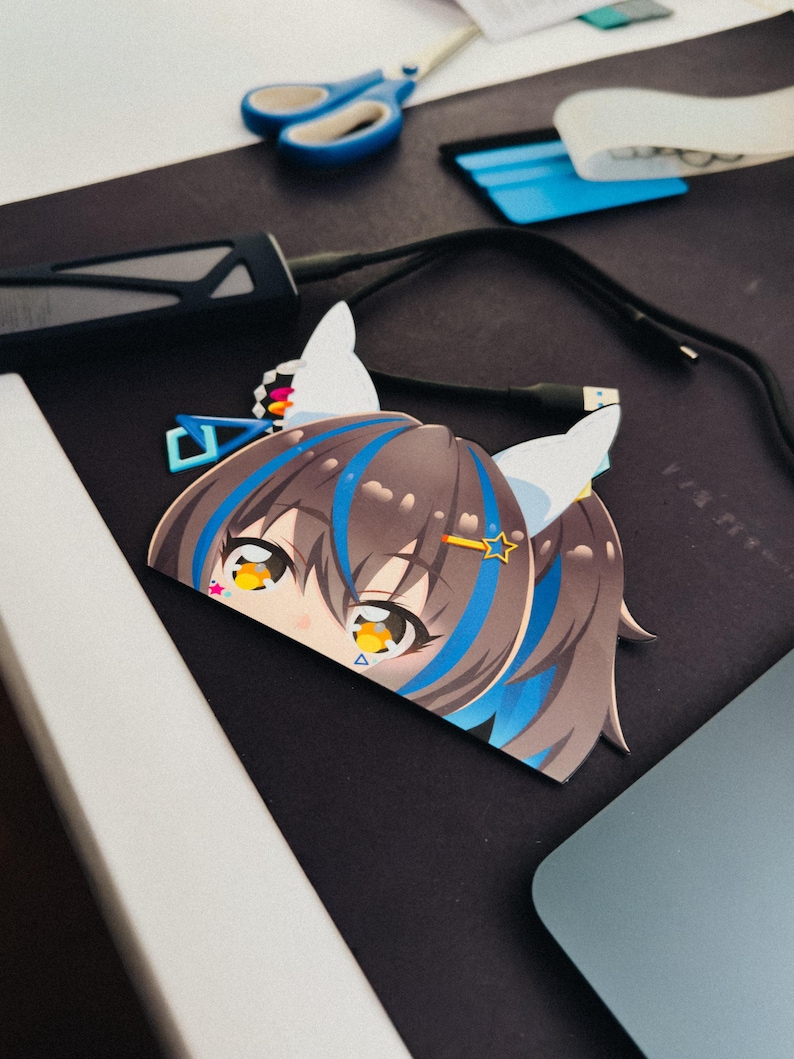 May include: A cut-out sticker featuring an anime-style character with brown hair, blue highlights, and large yellow eyes. The sticker has white cat ears and a star-shaped hair accessory. The background includes a black surface, scissors, and a blue tool.