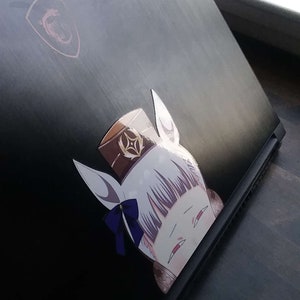 May include: Black laptop with a sticker of a cartoon character with white hair, a brown hat, and a blue bow. The laptop has a red logo in the upper left corner.