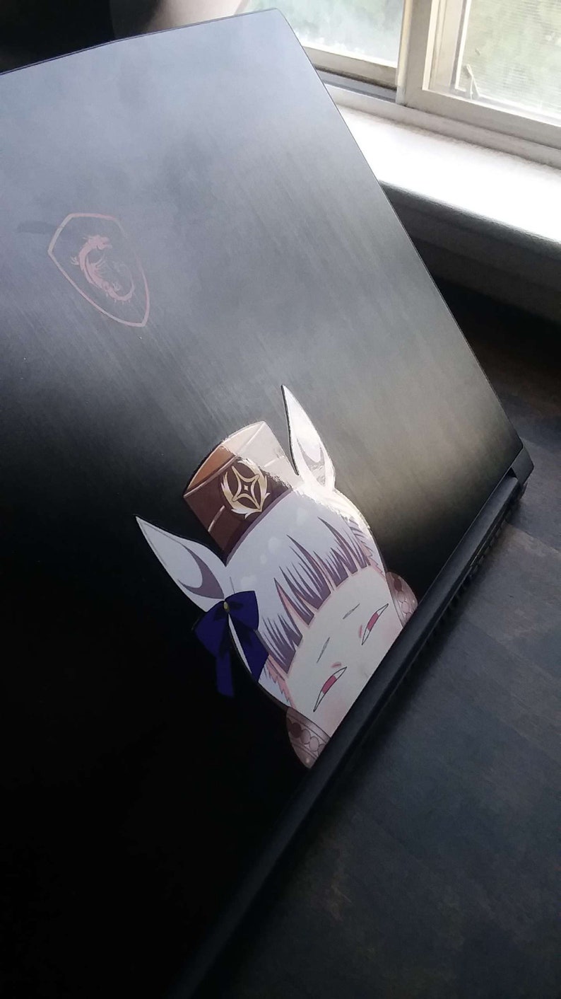 May include: Black laptop with a sticker of a cartoon character with white hair, a brown hat, and a blue bow. The laptop has a red logo in the upper left corner.