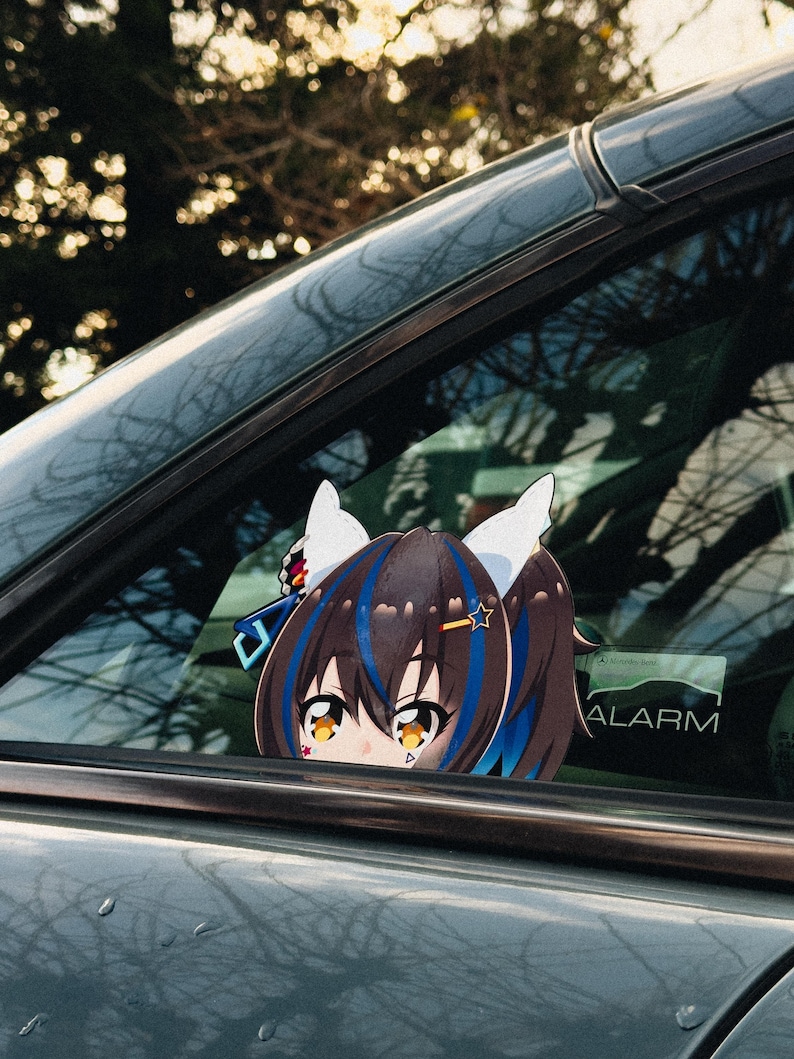 May include: A car window decal featuring an anime-style character with brown hair, blue highlights, and white ears. The decal is visible through the car window, with the word "ALARM" visible on the window.