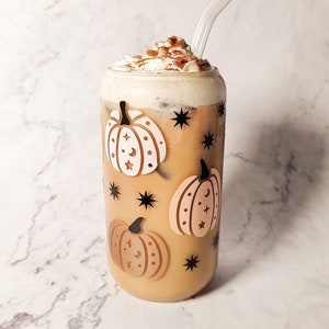 Peut inclure: Un verre de café glacé avec de la crème fouettée et de la cannelle, orné de motifs de citrouilles et d'étoiles. Le verre transparent est doté d'une paille incurvée. La boisson est posée sur un fond de marbre blanc.