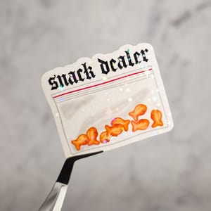 Pode incluir: Um adesivo branco com um saco de plástico transparente cheio de bolachas de peixe laranja. O adesivo tem o texto "snack dealer" em letras pretas.