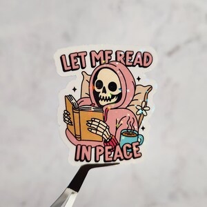 May include: A sticker featuring a cartoon skeleton in a pink hoodie reading a book while sitting on a pillow. The sticker has the text "LET ME READ IN PEACE" in pink and black. A cup of coffee and a flower are also included.