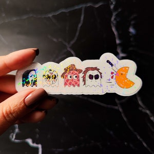 K&ouml;nnte beinhalten: Ein holografischer Aufkleber mit Cartoon-Versionen von Horrorfilmfiguren und einer Pac-Man-&auml;hnlichen Figur, die mit einem Messer erstochen wird. Zu den Figuren geh&ouml;ren Ghostface, Jason Voorhees, ein rosa Geist mit Cowboyhut und Michael Myers.