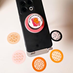 May include: A pink phone grip with a cartoon potato wearing a red armchair and the text "Just trying to get my chip together". The grip is attached to a black smartphone with a triple camera lens. There are four other phone grips in the background, each with a different pattern.