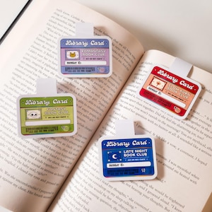 May include: Four colorful bookmarks with a library card theme. Each bookmark features a different book club theme: "Romantasy Book Club", "Fantasy Book Club", "Slow Burn Book Club", and "Late Night Book Club". Each bookmark has a cartoon illustration and a member ID number.