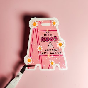 Puede incluir: Pegatina de señal de precaución rosa y blanca con purpurina y un diseño de flor sonriente. El letrero dice "No estoy de humor. Acércate con precaución."