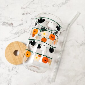May include: A clear glass tumbler with a bamboo lid and a clear straw. The glass is decorated with a string of orange pumpkins, white ghosts, and black bats. The tumbler is perfect for Halloween.