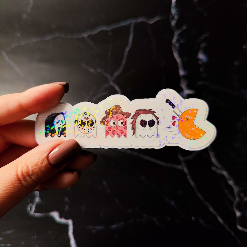 K&ouml;nnte beinhalten: Ein holografischer Aufkleber mit Cartoon-Versionen von Horrorfilmfiguren und einer Pac-Man-&auml;hnlichen Figur, die mit einem Messer erstochen wird. Zu den Figuren geh&ouml;ren Ghostface, Jason Voorhees, ein rosa Geist mit Cowboyhut und Michael Myers.