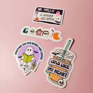 K&ouml;nnte beinhalten: Eine Sammlung von Aufklebern zum Thema Halloween auf rosa Hintergrund. Die Aufkleber zeigen gruselige Designs, darunter Geister, Flederm&auml;use und S&auml;tze wie "Hello A Spooky Ass Bitch" und "Ghosting You For Books and Coffee". Ein Aufkleber zeigt ein kaltes Getr&auml;nk mit dem Text "Cold Like My Heart".