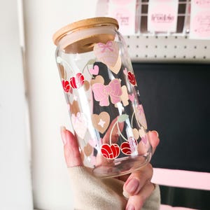 May include: A clear glass tumbler with a wooden lid, decorated with pink bows, hearts, and red cherries. The tumbler is held in a person's hand. The design is printed on the glass. The tumbler is designed for cold beverages.
